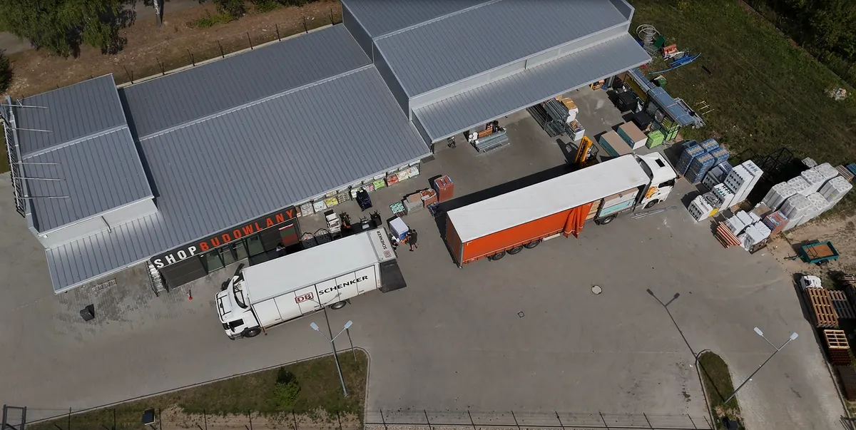 Skład SHOP BUDOWLANY z lotu ptaka — hala, plac manewrowy i ciężarówki podczas rozładunku