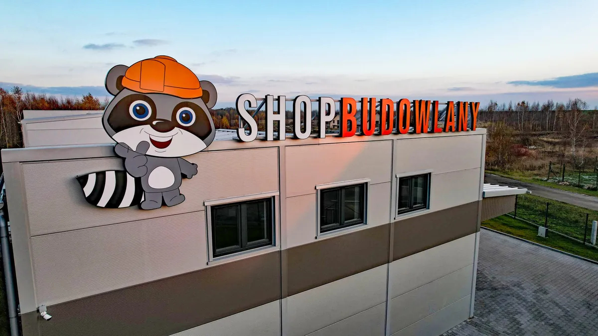 Szyld SHOP BUDOWLANY z maskotką szopa w kasku na dachu hali